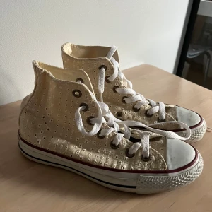 Beige Converse All Star broderade sneakers - Unika Converse All Star high tops i beige med broderat hålmönster och vita snören. Klassisk rund tå, vit gummisula med röd rand och Converse-logga på sidan. Använda fåtal gånger så i mycket bra skick fortfarande. Jättesöta nu till hösten eller våren för att piffa upp en tråkig outfit.