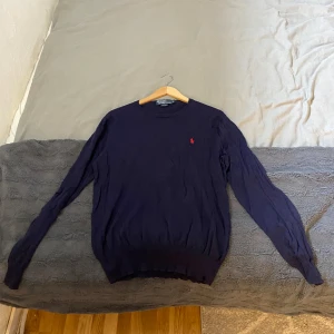 Mörkblå tröja från Polo Ralph Lauren - Säljer en mörkblå långärmad tröja från Polo Ralph Lauren i slim fit-modell. Tröjan har en klassisk rund halsringning och den ikoniska röda loggan broderad på bröstet. Perfekt för dig som gillar stilrena och tidlösa plagg.