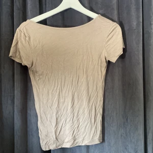 Beige topp med djup rygg från 157 - En enkel och stilren beige topp från 157 med korta ärmar och djup, rundad rygg. Toppen är i mjukt och stretchigt material som sitter snyggt på kroppen. Perfekt att matcha med jeans eller kjol för en clean look.