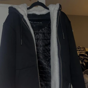 Moose Knuckles Classic Bunny  - Svart teddyjacka från Moose Knuckles med vit fluffig fuskpäls på insidan av huvan. Jackan har dragkedja framtill, snörning i huvan och två fickor. Insidan är quiltad med mönstrat foder och logga på ärmen. Perfekt för kalla dagar och riktigt snygg streetstil. Kvitto finns!