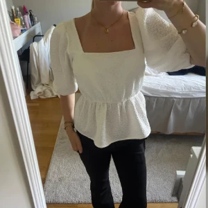 Vit blus med puffärm från Gina Tricot - Supersöt vit blus från Gina Tricot med puffiga trekvartsärmar och fyrkantig ringning. Blusen har en strukturerad, lite bubblig yta och markerad midja med volang nedtill. Perfekt att styla med jeans eller kjol.