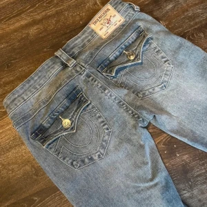 True Religion ljusblå bootcut jeans - Snygga ljusblå jeans från True Religion med bootcut passform och ikoniska bakfickor med lock och stora sömmar. Jeansen har klassisk femficksdesign, knappgylf och är tillverkade i mjukt denimtyg. Perfekta för dig som gillar en avslappnad men trendig look.