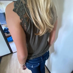 Olivgrön blus med spetsdetaljer - Snygg olivgrön blus med ärmlös design och rund hals. Axlarna har coola spetsdetaljer med blommönster som ger en extra touch. Blusen är lätt och skön, perfekt till jeans eller kjol. Materialet känns mjukt och luftigt, och modellen är normal i passningen.