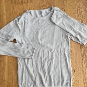 Ljusgrå långärmad tröja Jack & Jones - Snygg ljusgrå långärmad tröja från Jack & Jones i storlek M. Tröjan har en enkel och clean design med rund hals och diskret struktur i materialet. Perfekt för dig som gillar stilrena och bekväma plagg. Passar till jeans eller chinos.
