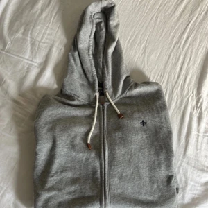 Grå  zip hoodie från Morris - Snygg grå hoodie från Morris  med dragkedja och huva. Tröjan har vita snören med bruna detaljer och en liten blå logga broderad på bröstet.  Den är använd cirka 10 gånger 