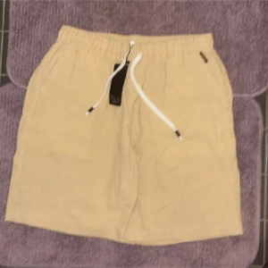 Beige shorts med snörning SPORT - Beiga shorts med elastisk midja och vit snörning framtill. Modellen har fickor på sidorna och en bakficka. Tillverkade i ett mjukt bomullstyg, perfekta för varma dagar. Enkel och clean design som passar till det mesta.