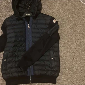 Svart Moncler pufferjacka med huva - Snygg svart pufferjacka från Moncler med quiltad framsida, huva och långa ärmar. Jackan har dragkedja framtill, fickor med tryckknappar och en extra ficka med dragkedja på ärmen. Perfekt för dig som vill ha en stilren och sportig look.