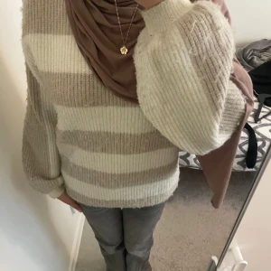 Beige och vit randig stickad tröja - Supermysig stickad tröja med breda beige och vita ränder. Tröjan har en relaxed fit med ballongärmar och ribbade muddar. Perfekt för dig som gillar en chill och stilren look under kyliga dagar.