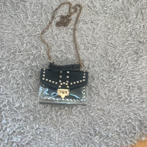 Svart axelväska med nitar och kedja - Cool axelväska i svart skinnimitation och transparent plast, dekorerad med guldiga nitar och lås. Väskan har en guldfärgad kedjerem och tydliga detaljer som ger en edgy vibe. Perfekt för dig som vill sticka ut med accessoarerna.