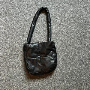 Svart quiltad axelväska i skinnimitation - Säljer en svart quiltad axelväska med vadderat handtag och rutmönstrad design. Väskan är i skinnimitation och har ett rymligt fack med dragkedja upptill. Perfekt storlek för mobilen, plånboken och lite till. Enkel och stilren look som passar till det mesta.
