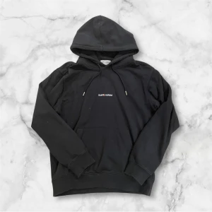 Svart hoodie från Saint Laurent - Svart hoodie från Saint Laurent med minimalistisk logga broderad på bröstet. Klassisk huva med snörning och känguruficka framtill. Tillverkad i mjuk bomull, perfekt för en clean och stilren look.