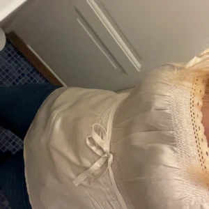 Söt guldig blus med spets och knyt - Romantisk gludig blus med korta ärmar, smock upptill och spetsdetaljer vid halsringningen. Midjan markeras med ett knytband för extra söt stil. Perfekt till jeans eller kjol för en fräsch look. Säljer i svart också 