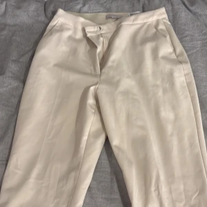 Beiga kostymbyxor från H&M, stl 38 - Snygga beiga kostymbyxor från H&M i storlek 38.Vida ben och klassisk passform med diskreta fickor bak och dragkedja fram. Perfekta för en clean och stilren look. Materialet är mjukt och känns bekvämt mot huden.