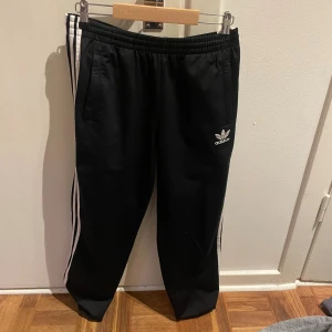 Svarta Adidas mjukisbyxor med vita ränder - Svarta mjukisbyxor från Adidas med klassiska vita ränder längs sidorna och broderad logga på låret. Byxorna har resår i midjan och raka ben, perfekta för chill eller träning. Materialet är mjukt och bekvämt.