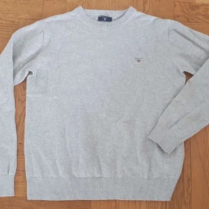 Ljusgrå bomullströja från Gant - En stilren ljusgrå tröja från Gant i mjuk bomull. Klassisk rund halsringning, ribbade muddar och diskret Gant-logga broderad på bröstet. Perfekt för dig som gillar enkel och clean stil.