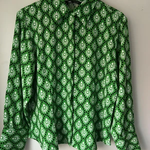 Grön blommig skjorta från Zara - Snygg grön skjorta från Zara med blommigt mönster i vitt och svarta prickar. Skjortan har klassisk krage, knäppning framtill och långa ärmar. Perfekt för dig som vill sticka ut med färg och mönster.