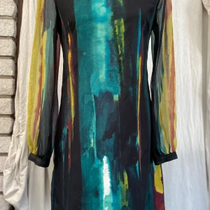 Färgstark långärmad klänning Indiska S - Färgglad klänning med långa, genomskinliga ärmar. Klänningen har ett abstrakt mönster i nyanser av svart, grönt, gult och rött. Den har en rund halsringning och en rak passform. Tvättråd saknas! ﻿Längd: 87 cm ﻿Bystvidd: 47 cm  Inga bilder när den sitter på mig! Katter finns i hemmet! 01,S