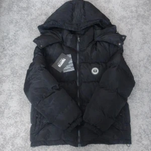 Trapstar jacka - Svart pufferjacka från Trapstar med stor huva och dragkedja framtill. Jackan har broderad Trapstar-logga på ryggen och en rund patch på bröstet. Perfekt för kalla dagar. Snygg med tydlig branding.