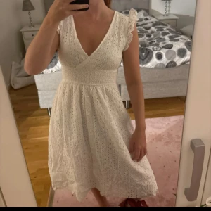 Vit omlottklänning med volangärm - Superfin vit klänning i bomull med broderade detaljer och omlott-v-ringning. Klänningen har korta volangärmar, smock i midjan och ett luftigt, lätt tyg. Perfekt för sommarens alla tillfällen och ger en romantisk vibe.