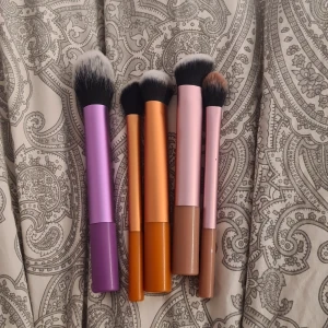 Set med sminkborstar i färgmix - Säljer ett set med fem sminkborstar från Real Techniques. Borstarna har handtag i lila, orange, rosa och beige och mjuka syntetiska borst i svart och vitt. Perfekta för applicering av foundation, rouge, highlighter och contour. Snygg design och sköna att använda.