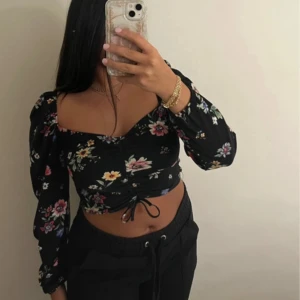 Svart blommig croppad blus med snörning - Snygg svart blus med färgglatt blommönster, croppad passform och v-ringad hals. Toppen har långa puffärmar och snörning framtill som ger en trendig look. 