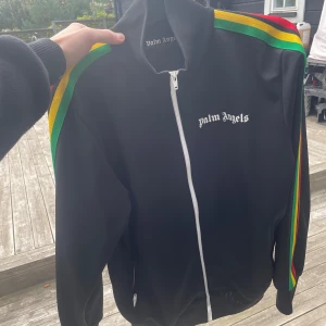 Palm Angels svart track jacket L - Palm Angels svart track jacket med vita dragkedjor och logga på bröstet. Ärmarna har färgglada ränder i grönt, gult och rött för en riktigt clean streetstyle-look. Jackan är gjord i 100% polyester och har hög krage och fickor med dragkedja.