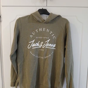 Olivgrön hoodie från Jack & Jones - Olivgrön hoodie från Jack & Jones med vit tryckt logga på bröstet. Tröjan har huva, långa ärmar och ribbade muddar. Tillverkad i mjuk bomullsmix som känns skön mot huden. Perfekt för en avslappnad och stilren look. En stor fläck inne i luvan, som man dock inte ser, defrö billigt pris