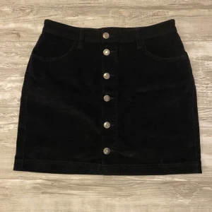 Svart kjol med knappar - Snygg svart kortkjol med fickor fram och hällor för bälte, perfekt för en cool och enkel stil. Kjolen är i storlek 10 och passar som storlek S