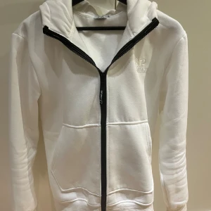 Vit hoodie med goggles från C.P. Company - Vit hoodie från C.P. Company med ikoniska goggles i huvan och broderad logga på bröstet. Tröjan har hel dragkedja framtill, känguruficka och är tillverkad i mjuk bomull. Perfekt för dig som vill sticka ut med en streetwear vibe.