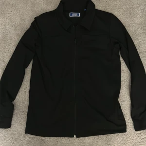 Svart overshirt från Jack & Jones - Stilren svart overshirt från Jack & Jones med klassisk krage och hel dragkedja framtill. Jackan har lång ärm och en minimalistisk look som passar perfekt till lager-på-lager. Materialet känns mjukt och smidigt, vilket gör den enkel att bära hela dagen.