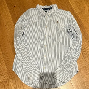 Randig skjorta från Ralph Lauren - Snygg skjorta från Ralph Lauren i vitt och blått med tunna vertikala ränder. Klassisk krage, långa ärmar och knäppning framtill. Broderad logga på bröstet. Perfekt för en clean och stilren look.