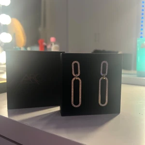 Glittriga guldörhängen från AROCK - Snygga statement-örhängen i guld från AROCK med rektangulär länkdesign. Örhängena är dekorerade med glittrande stenar längs hela kanten för extra bling. Perfekta för att lyfta din look med lite extra glamour.