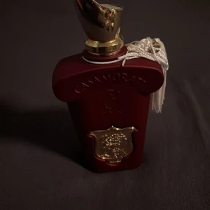 Casamorati Eau de Parfum 100ml med 95% kvar i flaskan  - Lyxig parfymflaska från Casamorati i matt vinröd färg med gulddetaljer och vit tofs. Flaskan har en unik form med elegant guldlock och dekorativt emblem framtill. Rymmer 100ml Eau de Parfum och utstrålar exklusivitet och vintagekänsla.