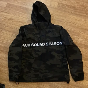 Camo vindjacka från Black Squad S - Snygg camouflagemönstrad vindjacka från Black Squad i storlek S. Jackan har huva med dragsko, halv dragkedja framtill, stor ficka på ärmen och tryckt text över bröstet. Perfekt för dig som gillar streetwear och vill sticka ut.