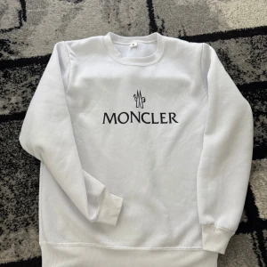 Moncler tröja - Säljer en clean vit sweatshirt från Moncler med svart logga tryckt på bröstet. Tröjan har rund halsringning, långa ärmar och ribbade muddar. Perfekt för dig som gillar stilrena och klassiska plagg med premiumkänsla.