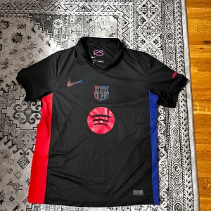 FC Barcelona svart matchtröja Nike - Snygg FC Barcelona fotbollströja från Nike i svart med röda och blå sidopaneler. Tröjan har korta ärmar, v-ringad krage och detaljer som klubbmärke, Nike-logga och Spotify-tryck på bröstet. Materialet är lätt och andas, perfekt för träning eller match.