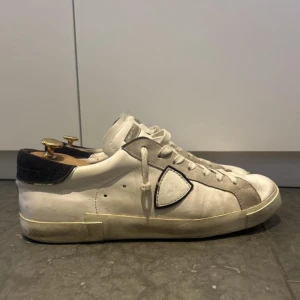 Vita sneakers från Philippe Model - Snygga vita sneakers från Philippe Model med grå mockadetaljer och svart häl. Klassisk låg modell med snörning och rund tå. Skorna har en ikonisk sköldlogga på sidan och platt sula. Perfekta för en stilren och avslappnad look.