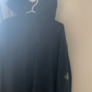 Svart hoodie från Calvin Klein Jeans - Svart hoodie från Calvin Klein Jeans med klassisk huva och snörning. Tröjan har lång ärm och en tydlig CK-logga på ärmen. Mjuk och skön, perfekt för chill dagar. Snygg och enkel design som funkar till det mesta.