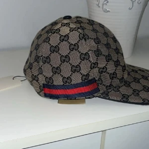 Gucci beige keps med GG-mönster - Snygg beige keps från Gucci med klassiskt GG-monogram i svart över hela kepsen. Har en justerbar rem i mörkblått skinn och detaljer med röd och blå rand på sidan. Materialet är bomull och polyester, insidan är mörkblå. Perfekt för dig som gillar lyxiga accessoarer.