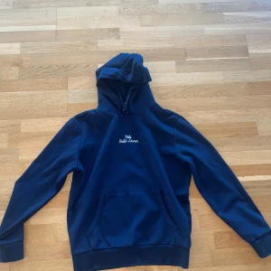 Mörkblå hoodie från Polo Ralph Lauren - Mörkblå hoodie från Polo Ralph Lauren med broderad vit logga på bröstet. Klassisk känguruficka framtill, huva med snörning och långa ärmar. Skön och stilren tröja i bomull, perfekt för chill dagar.