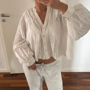 Vit blus med spets och vida ärmar - Säljer en luftig vit blus i bomull med v-ringning, knappar framtill och breda, ballongformade ärmar. Blusen har fina spetsdetaljer längs knappraden och axlarna, samt breda muddar vid ärmsluten. Perfekt för en chill och trendig look.