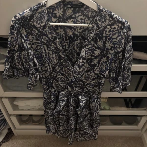 Blåvit omlottklänning från Zara - Supersöt kort omlottklänning från Zara i blått och vitt med blommigt mönster. Klänningen har volangdetaljer, v-ringning och korta ärmar. Perfekt för sommarens alla tillfällen och ger en fräsch och trendig look.