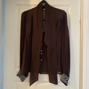 Brun blus från Wera med knappar - Säljer en brun, tunn och flowy blus från Wera i storlek 38. Skjortan har långa ärmar, knappar framtill och coola detaljer på manschetterna. Perfekt för dig som gillar en lite mer dressad men chill stil.