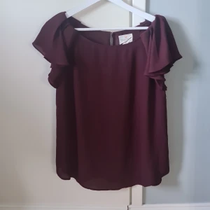 Vinröd blus från Holly & Whyte - Vinröd blus från Holly & Whyte by Lindex i luftig polyester. Blusen har korta volangärmar och en rund halsringning. Baksidan har en liten guldig knappdetalj vid nacken. Perfekt för dig som gillar en enkel men elegant stil.