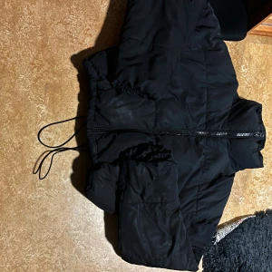Svart pufferjacka med dragsko - Säljer en svart pufferjacka med dragkedja och dragsko i midjan. Jackan har hög krage och långa ärmar, perfekt för kalla dagar. Enkel och stilren design som funkar till det mesta. Materialet är syntet och jackan är quiltad för extra värme.