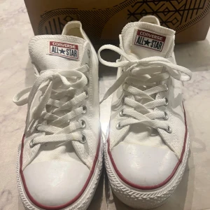 Vita Converse All Star sneakers - Klassiska vita Converse All Star sneakers i canvas med lågt skaft. Skorna har vit ovandel, vita snören, rund tå och platt sula med röd och blå rand längs sulkanten. Perfekta för en avslappnad och cool stil. Levereras i originalkartong.