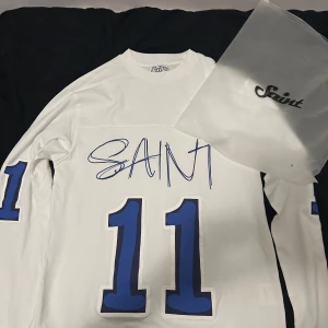 Vit långärmad Saint tröja  - Vit långärmad fotbollströja från Saint med stora blå nummer 11 på bröstet och ärmarna. Snyggt tryck med Saint-loggan framtill. Tröjan är i mjukt bomullsmaterial och har en klassisk passform med rund hals.