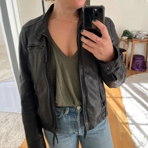 Svart skinnjacka med dragkedjor - Snygg svart skinnjacka med dragkedjor framtill och på fickorna. Jackan har en klassisk krage, lång ärm och en figurnära passform. Perfekt för dig som vill ha en tidlös och cool look till jeans eller klänning.