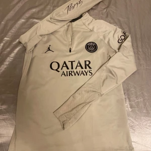 PSG långärmad träningströja Jordan - Snygg beige långärmad PSG träningströja från Jordan med svart Paris Saint-Germain-logga, Jordan-logga och Qatar Airways-tryck på bröstet. Dragkedja vid halsen, tryck på ärmen och Visit Rwanda-tryck på ryggen. Tillverkad i mjukt funktionsmaterial.