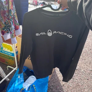 Svart långärmad tröja Sail Racing - Svart långärmad tröja från Sail Racing med vit logga tryckt framtill och en mindre logga i nacken. Klassisk rund halsringning och stilren design, perfekt för dig som gillar sportiga och cleana plagg.